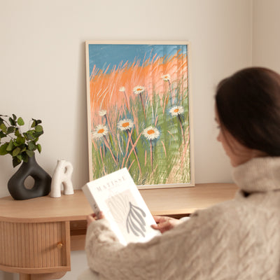 Person holding a book in a room with a painting of flowers on the wall.